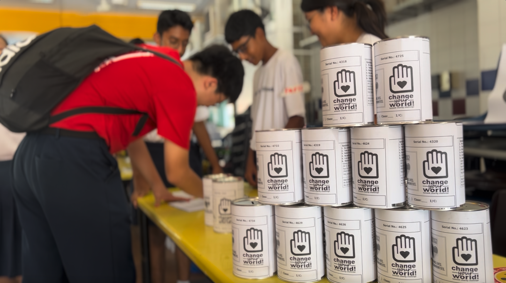 Students stand near a table stacked with white charity donation tins labeled "change your world" with a hand and heart symbol.